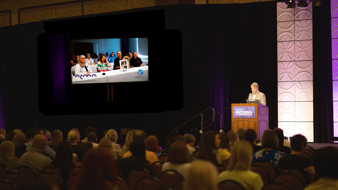 Speaker presenting at National Conference podium with audience and panel on overhead screen