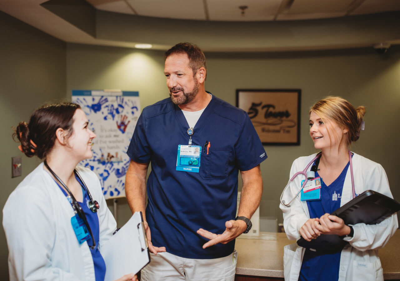 Three healthcare professionals wearing ID badges discuss patient care in a hospital corridor.