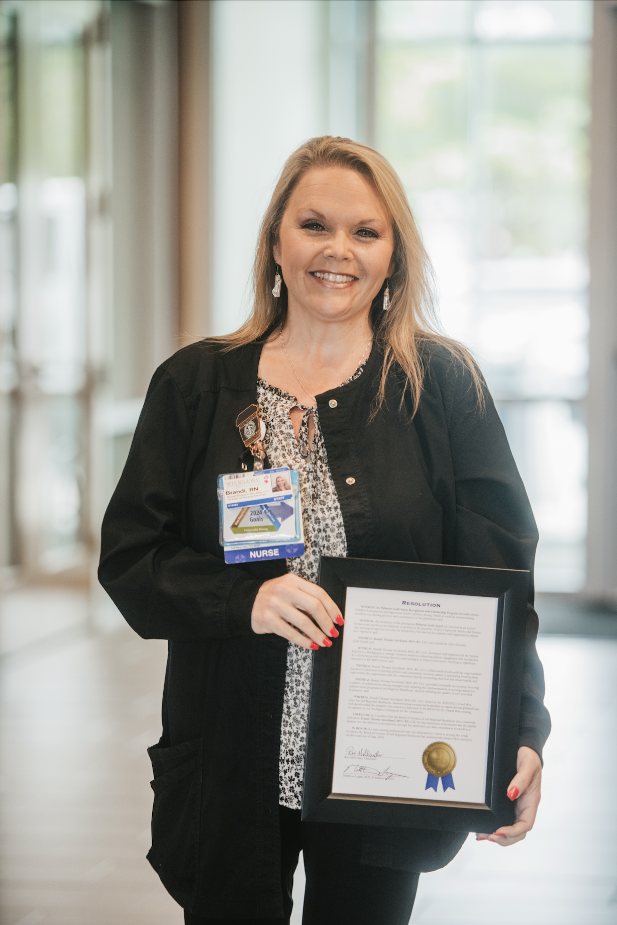 Healthcare professional holding a framed certificate and employee badge in a hospital corridor.