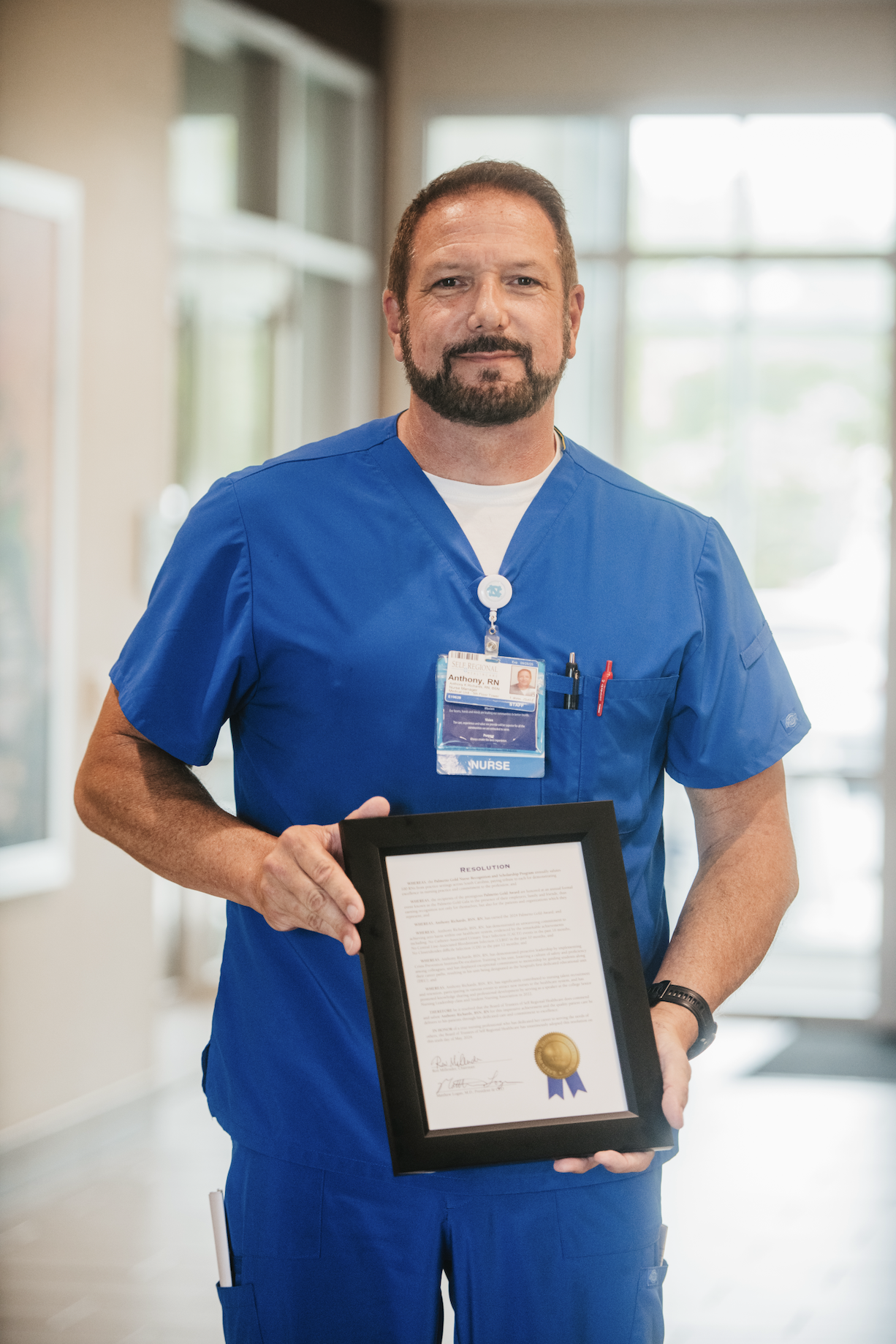 Healthcare professional in blue scrubs holding framed recognition award in hospital hallway.