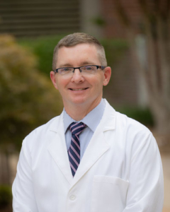 Healthcare provider in white coat and glasses smiling at camera outdoors