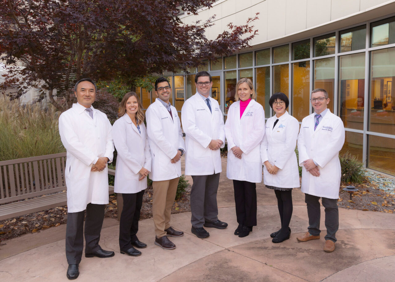 Seven healthcare professionals in white coats standing together outside a medical facility building.