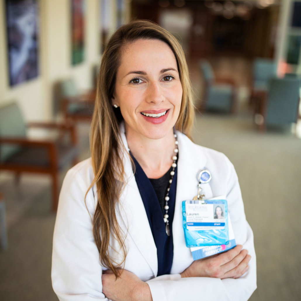 Healthcare professional wearing white coat and ID badge, smiling at camera in hospital corridor.