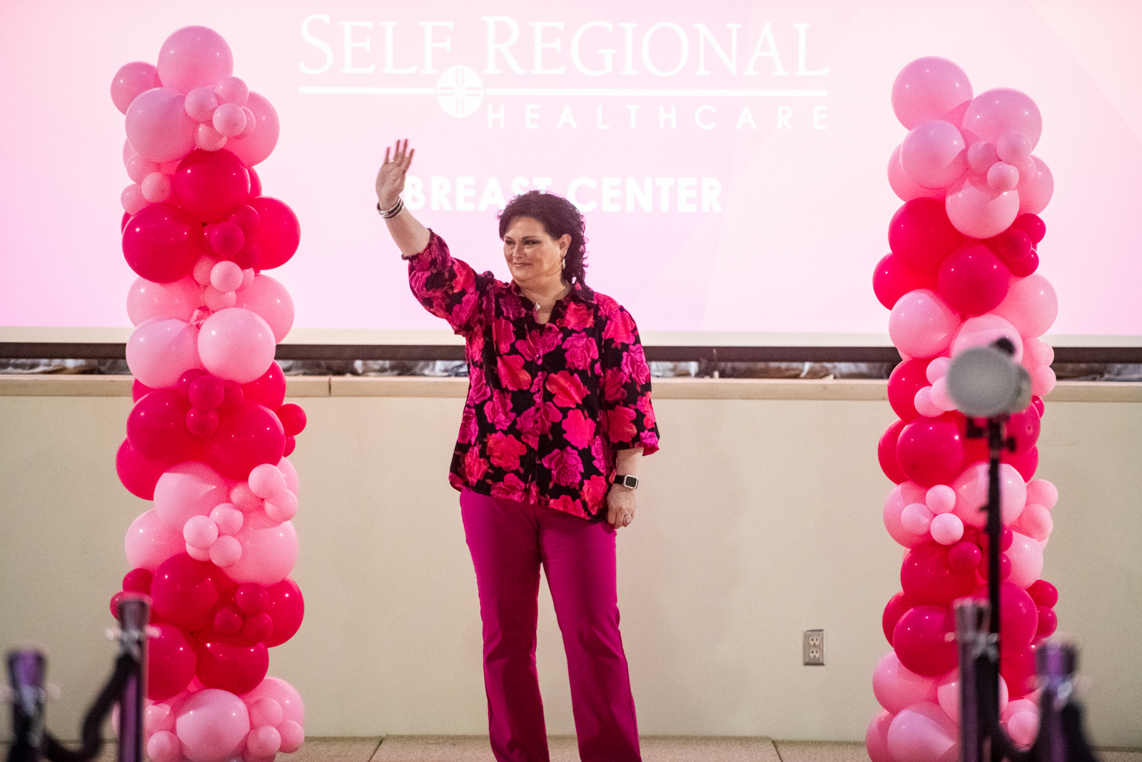 Woman waving at Self Regional Healthcare event with pink balloon columns flanking the stage