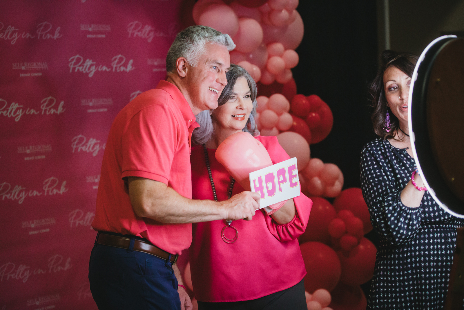 Man and woman holding "HOPE" sign at pink-themed healthcare event with balloon backdrop.
