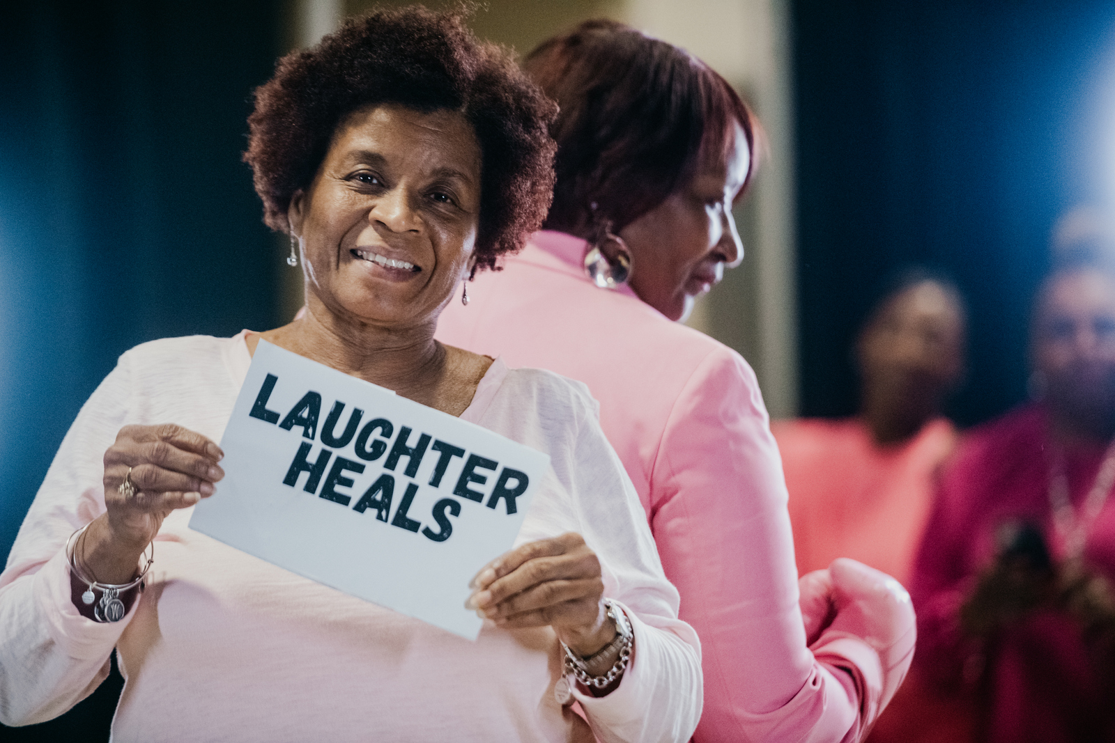 Woman holding "Laughter Heals" sign at healthcare wellness event with participants in background.