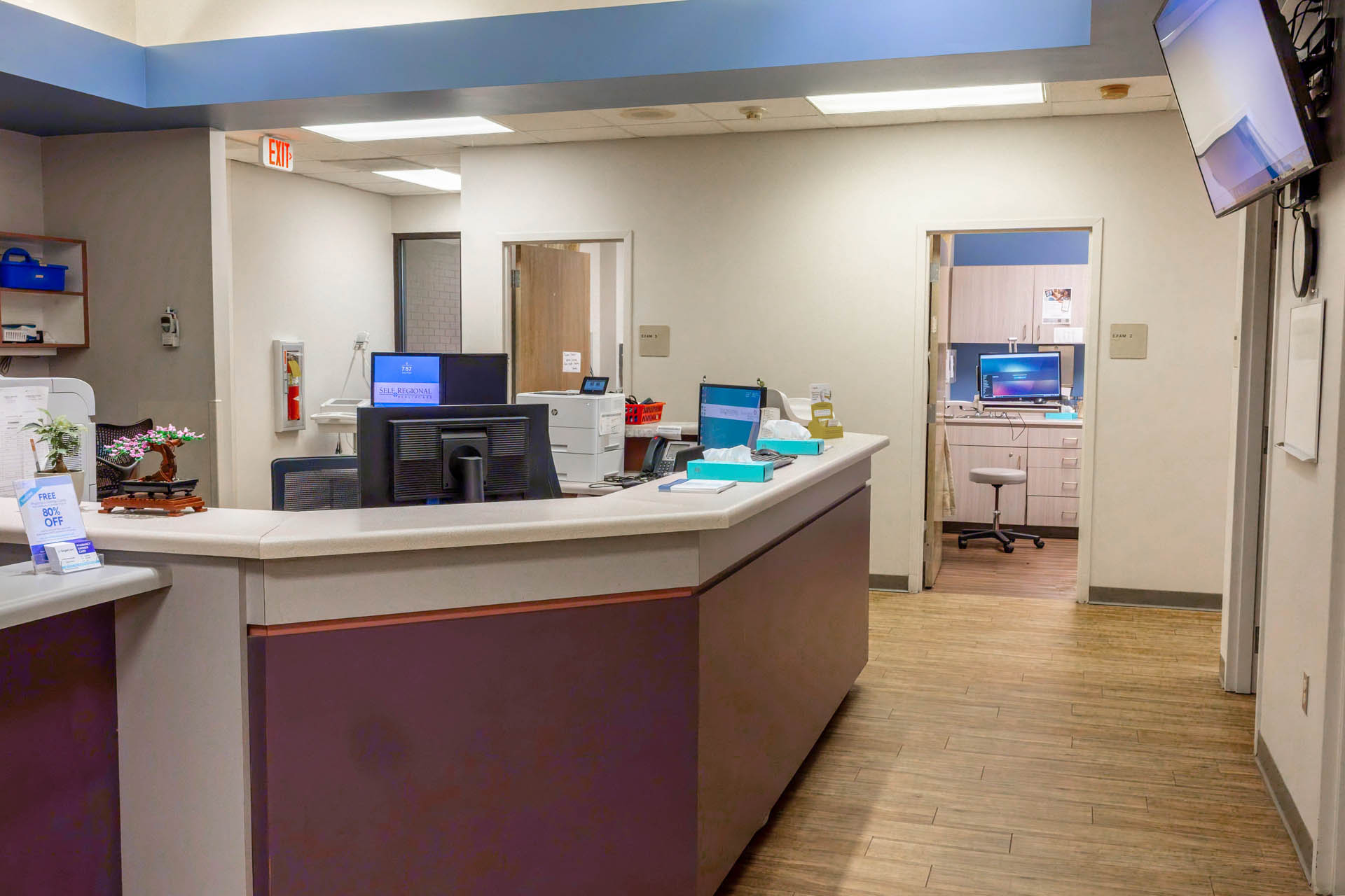 Healthcare clinic reception area with check-in desk, computers, and waiting room entrance.