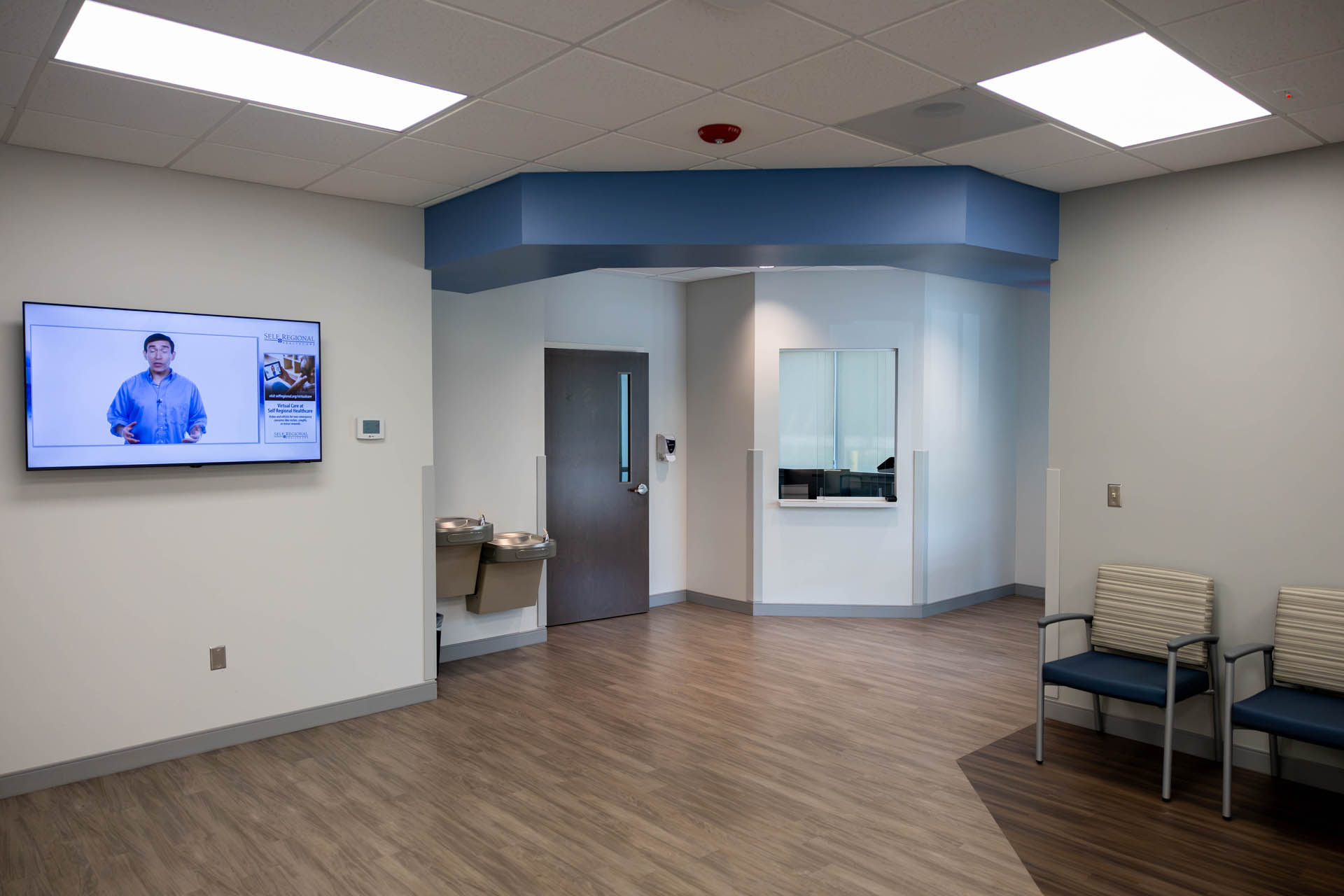 Urgent care waiting area with wall-mounted television, water fountains, reception window, and patient seating