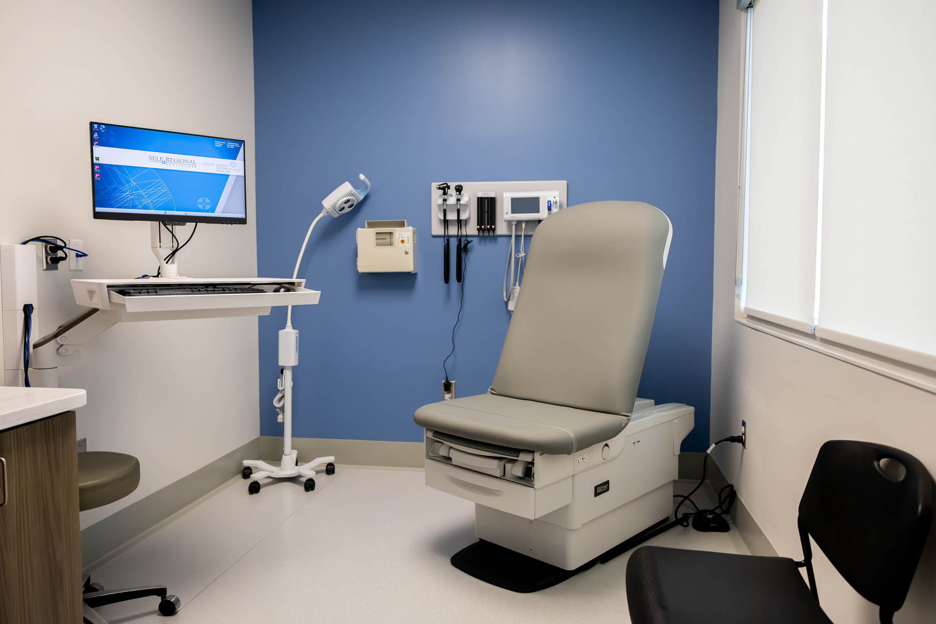Examination room with exam chair, computer workstation, and wall-mounted diagnostic equipment